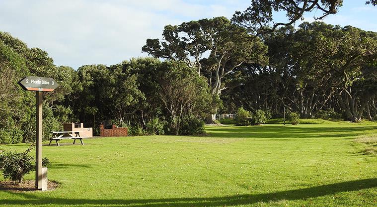 Wenderholm Regional Park - Bookable site three sign with open space, picnic table and trees in the background.