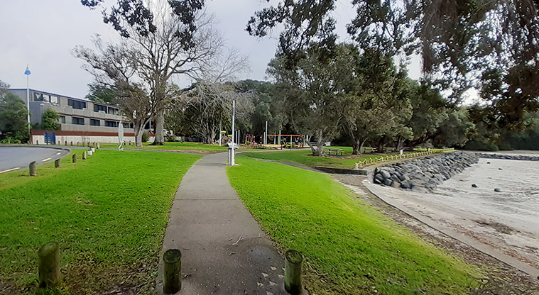 Cockle Bay Reserve - Section of path with trees and the playground in the background, and a section of the seawall and beach on the right.